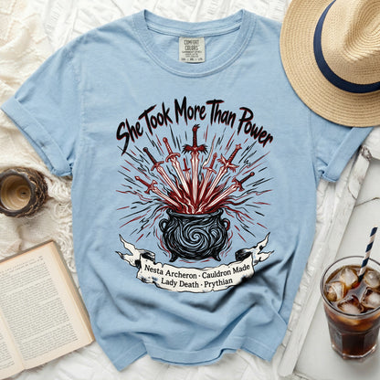 Light blue t-shirt with "She Took More Than Power" text, swords, and cauldron graphic.