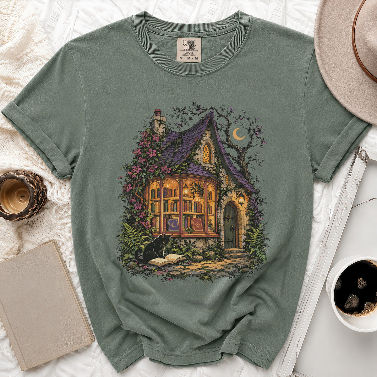 T-shirt with a witch's cottage illustration, a black cat reading, and a crescent moon.