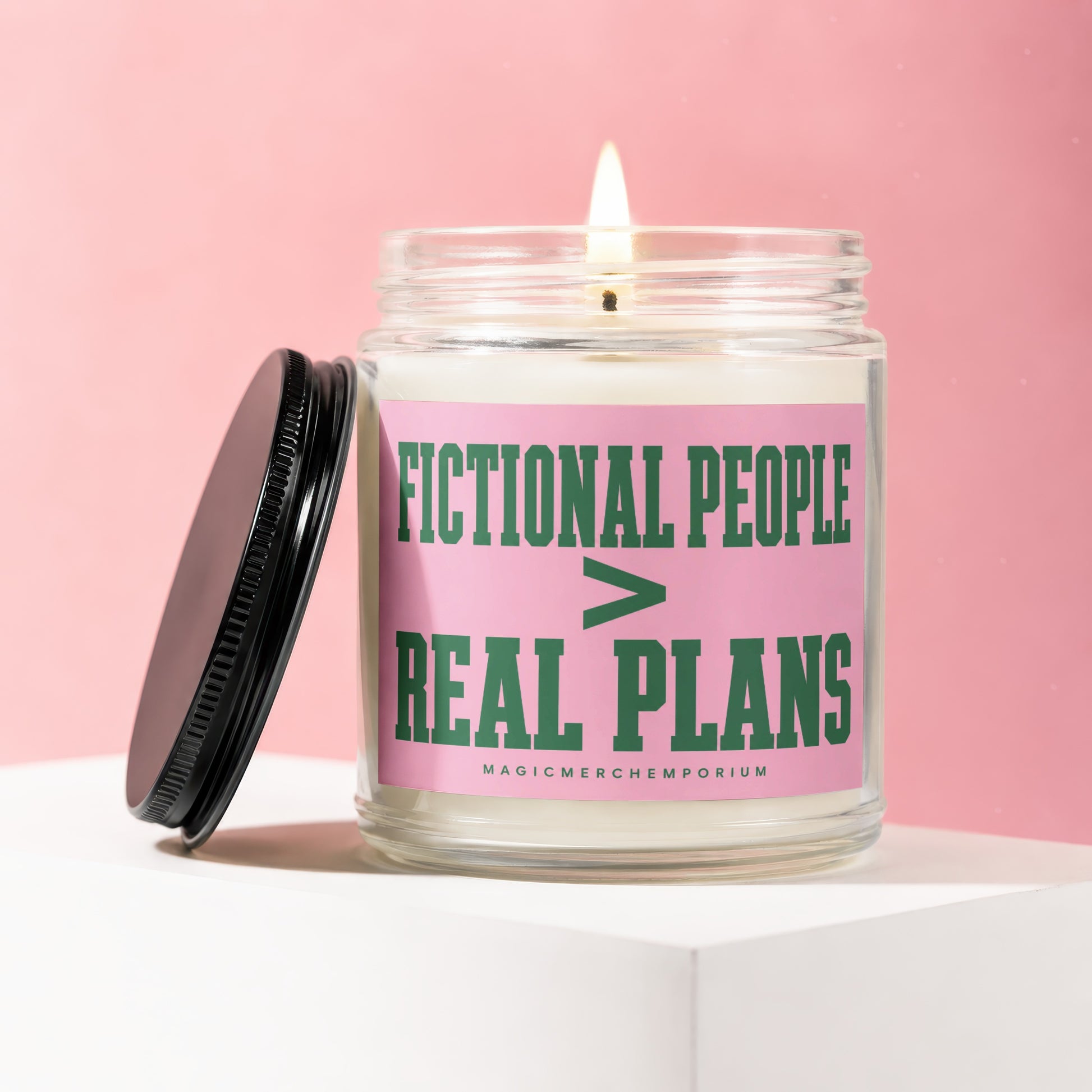 Glass candle with lit flame and black lid, featuring pink label with green text.