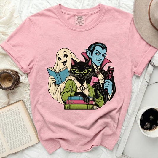 Pink t-shirt with graphic of ghost, black cat, and vampire reading books.