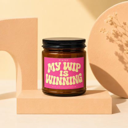 Amber jar candle with pink label reading "STATUS: MY WIP IS WINNING".