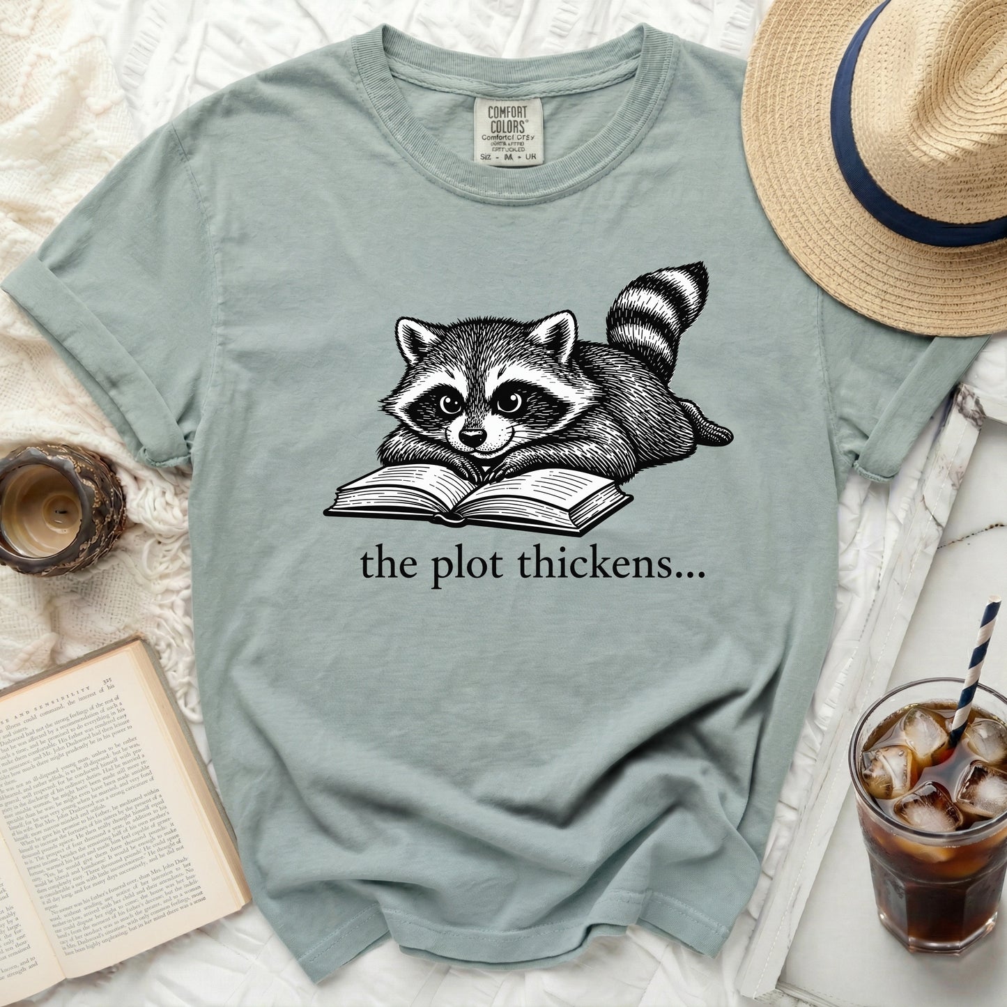 Green t-shirt with raccoon reading a book, "the plot thickens..." text
