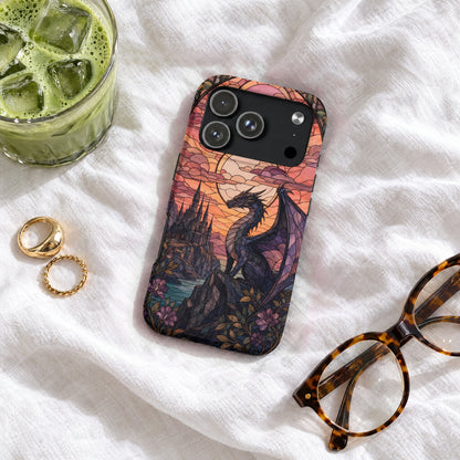 A smartphone case with a stained-glass dragon and castle design rests on white fabric next to glasses, rings, and a drink.