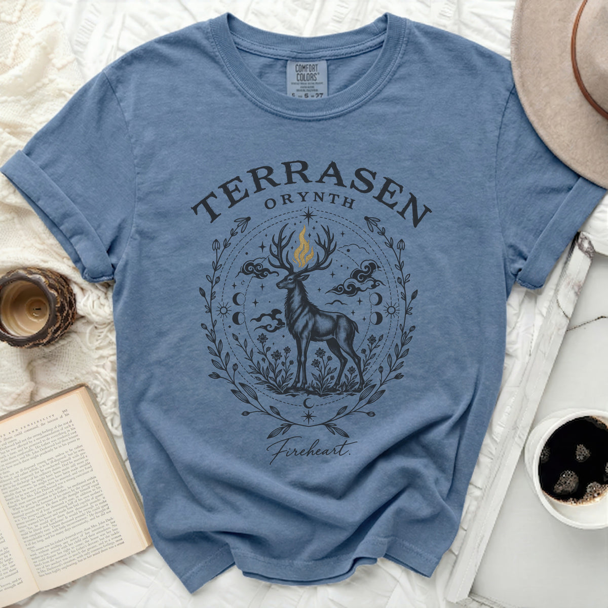 A blue crewneck t-shirt featuring a fantasy graphic of a stag with a flaming antler, surrounded by a floral and celestial border, with text reading Terrasen, Orynth, and Fireheart.