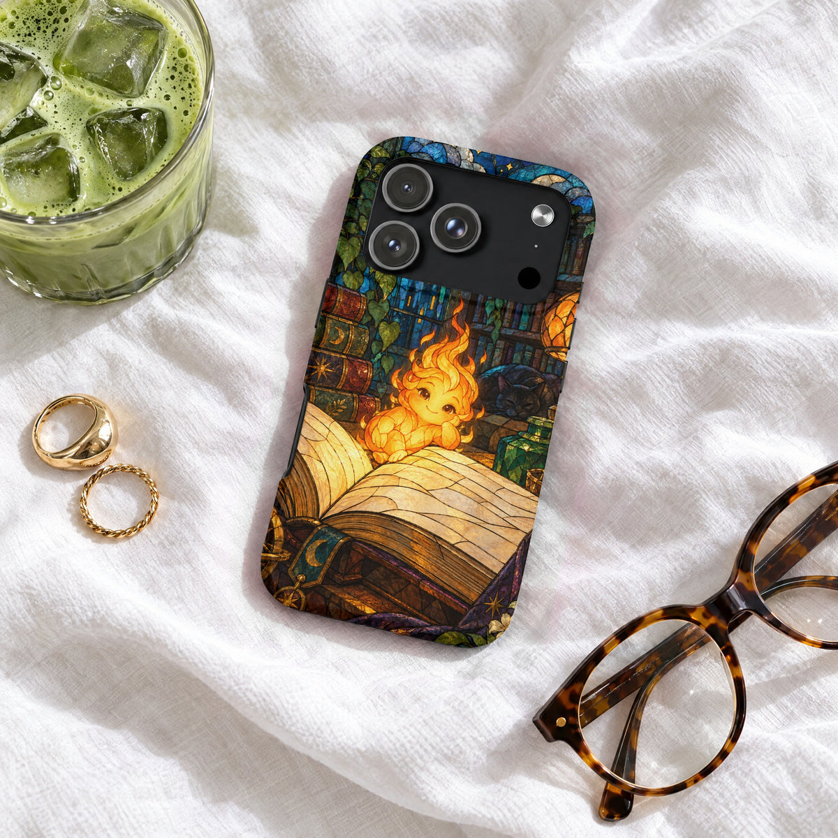 An iPhone case featuring a stained glass design of a fire sprite reading a book, resting on a white blanket with glasses, rings, and a glass of iced matcha.