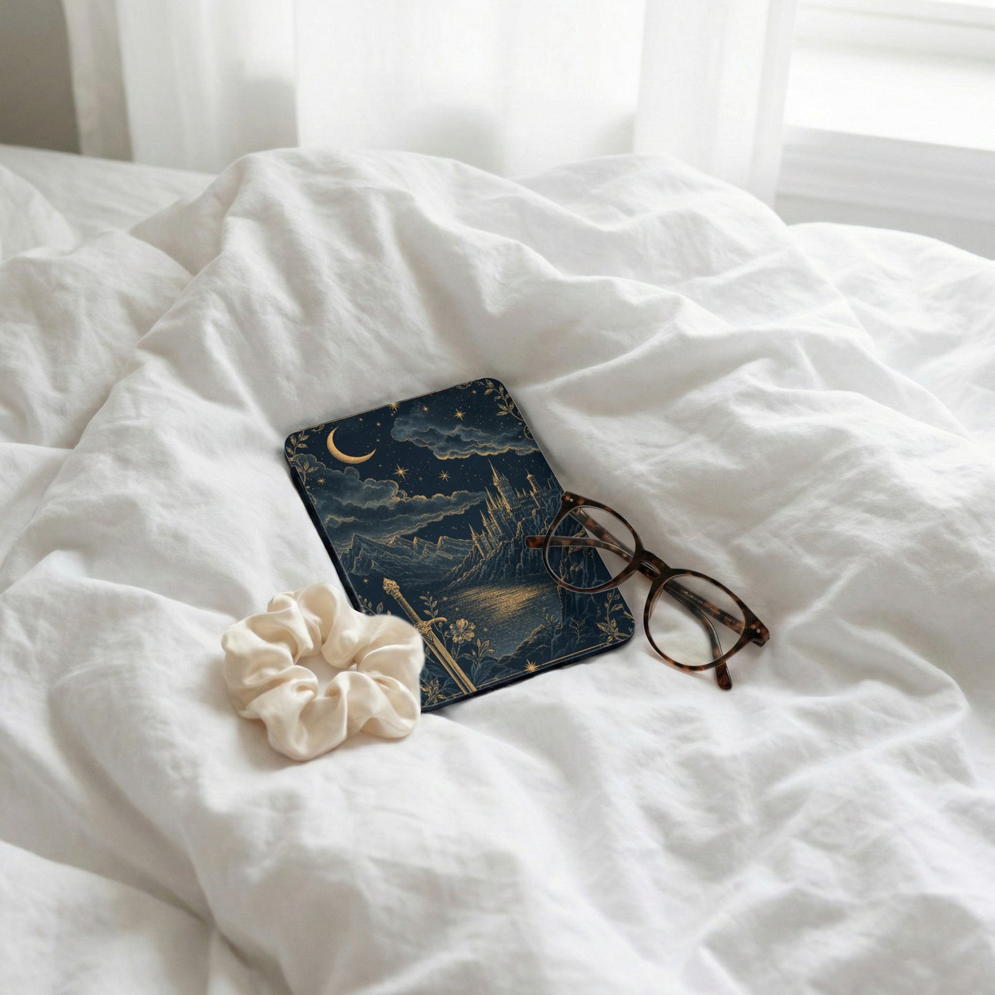 Book with celestial castle design, eyeglasses, and scrunchie on white bedding.