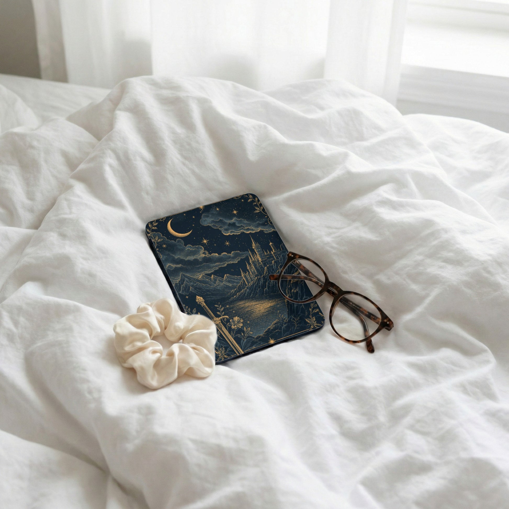 Book with celestial castle design, eyeglasses, and scrunchie on white bedding.