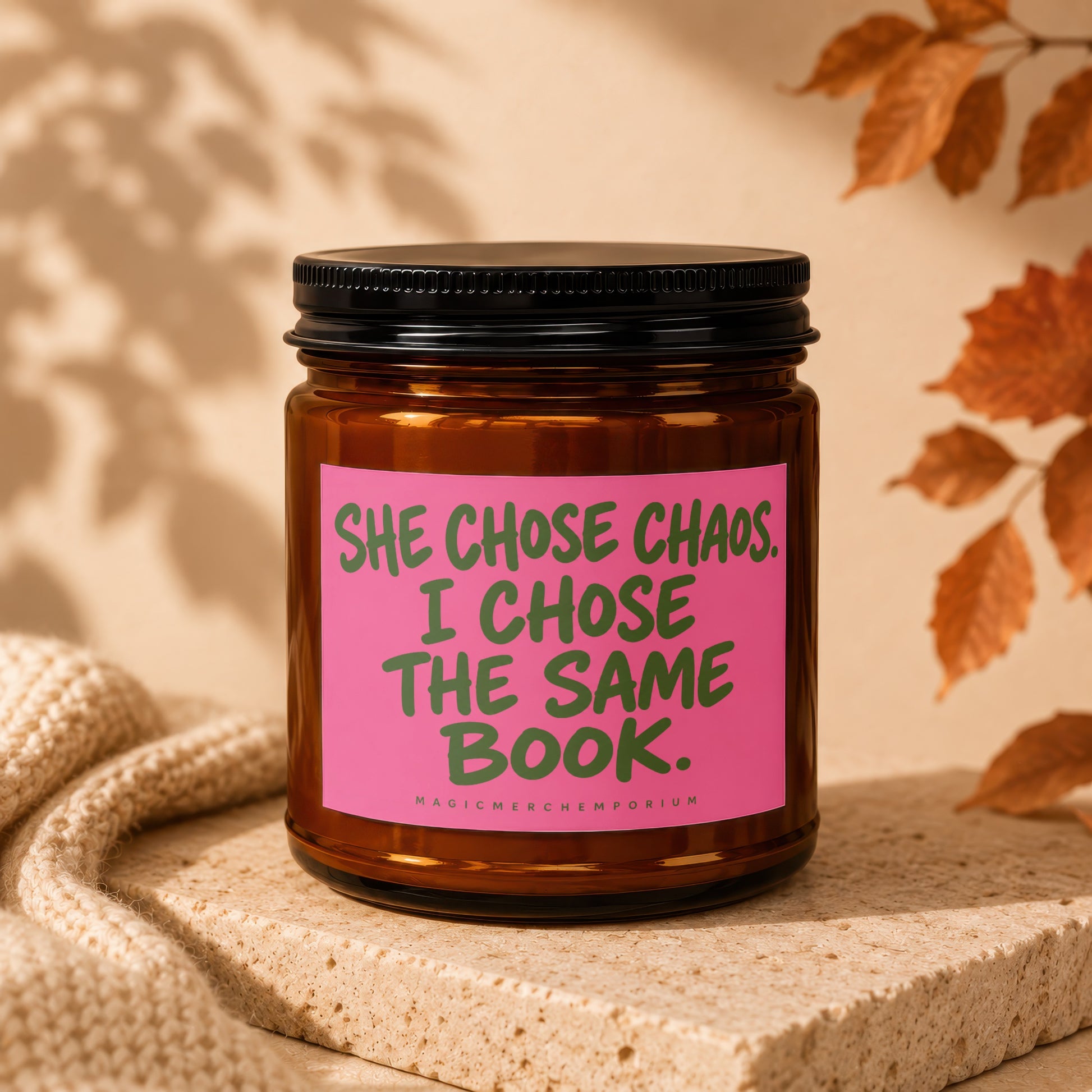 Amber jar candle with pink label reading "She chose chaos. I chose the same book."