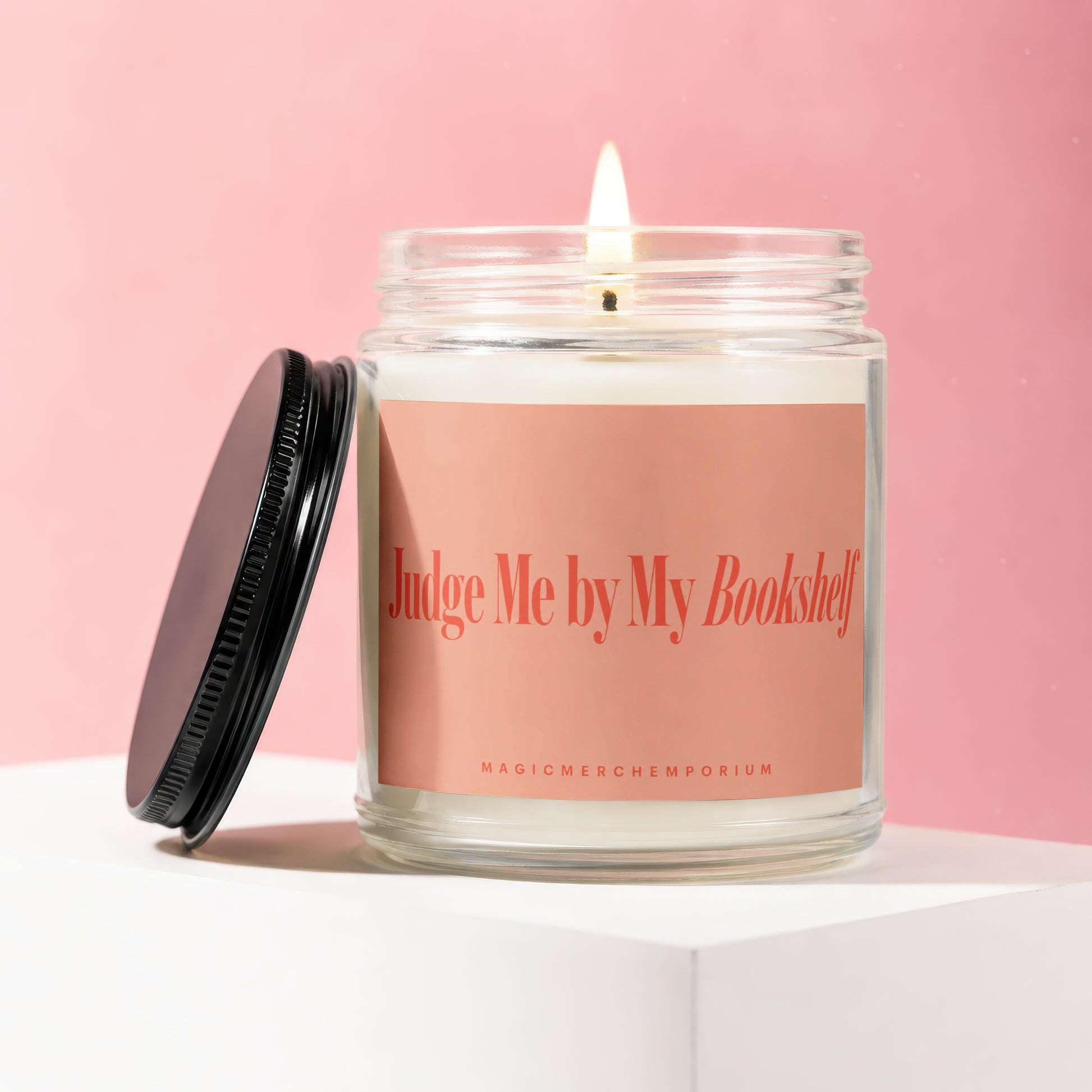 Glass jar candle with lit wick, reads "Judge Me by My Bookshelf."