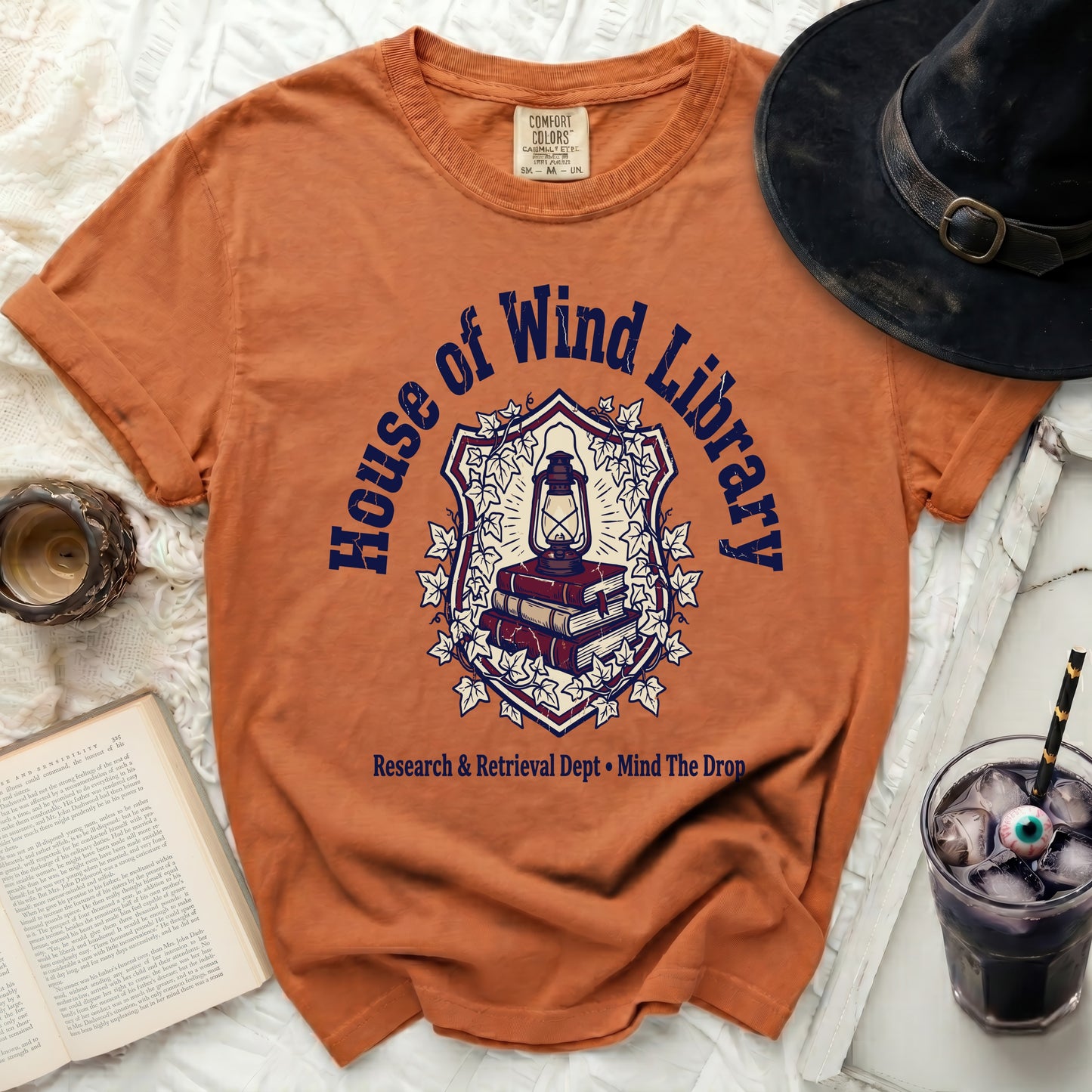 Orange t-shirt with "House of Wind Library" shield graphic and text.