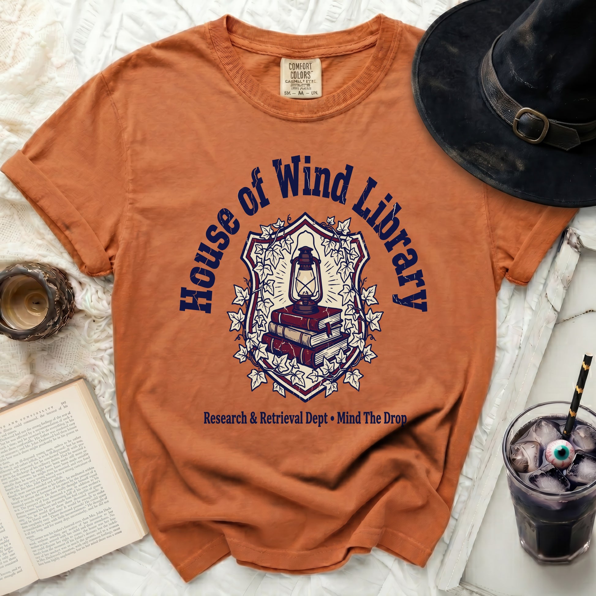 Orange t-shirt with "House of Wind Library" shield graphic and text.