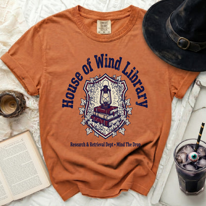 Orange t-shirt with "House of Wind Library" shield graphic and text.