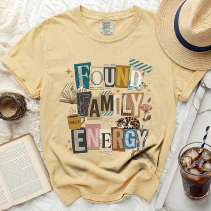 Yellow t-shirt with "FOUND FAMILY ENERGY" collage design, book, coffee, and cat.