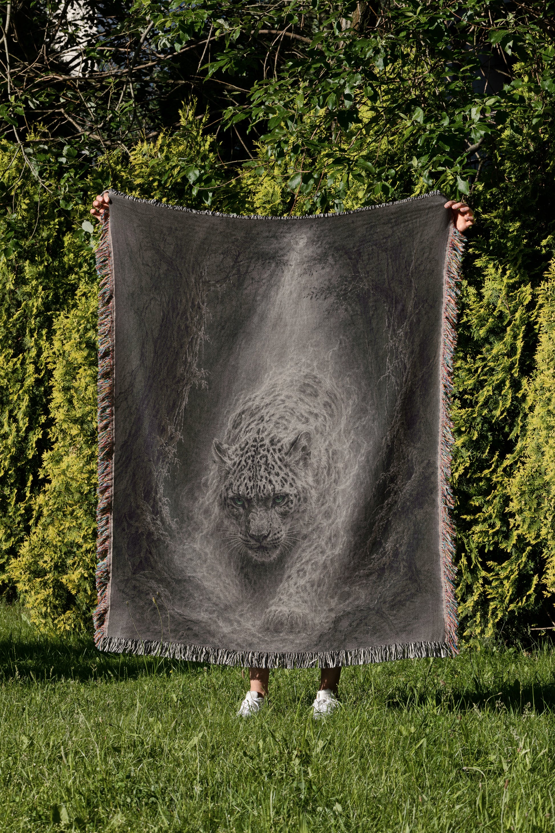 A person is standing in a grassy area, holding up a large black and white blanket that features a detailed image of a leopard's face.