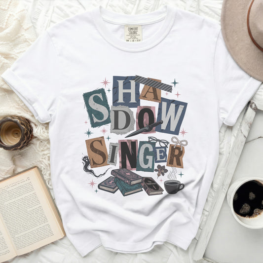 White t-shirt with "SHADOW SINGER" collage graphic, books, and glasses.