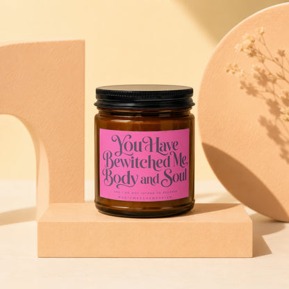 Amber glass candle with pink label reading "You Have Bewitched Me, Body and Soul."