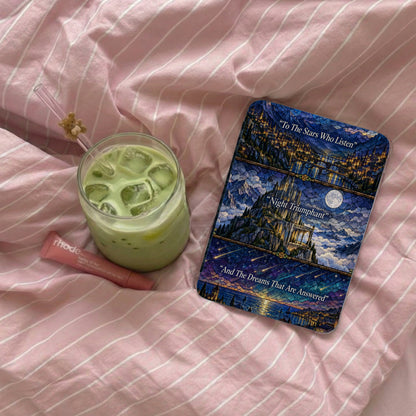 An e-reader with a fantasy stained-glass case, an iced matcha drink, and lip gloss on pink striped bedding.