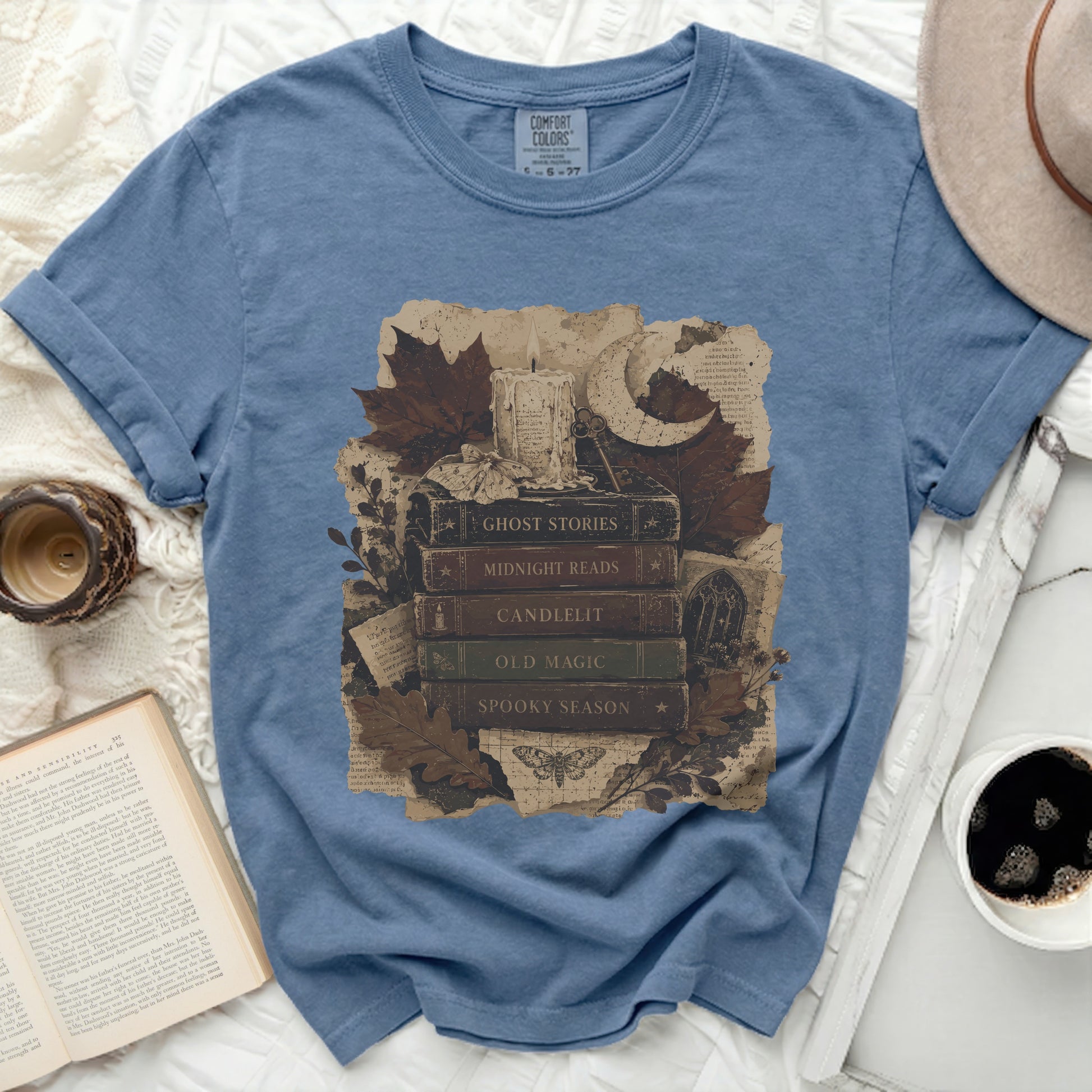 Blue t-shirt graphic with stack of books, candle, moon, and autumn leaves.