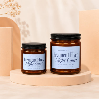 Two amber glass candle jars with black lids and light blue labels reading Frequent Flyer, Night Court stand on a pedestal.