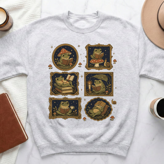 Grey sweatshirt with six framed illustrations of frogs reading books among mushrooms and stars.