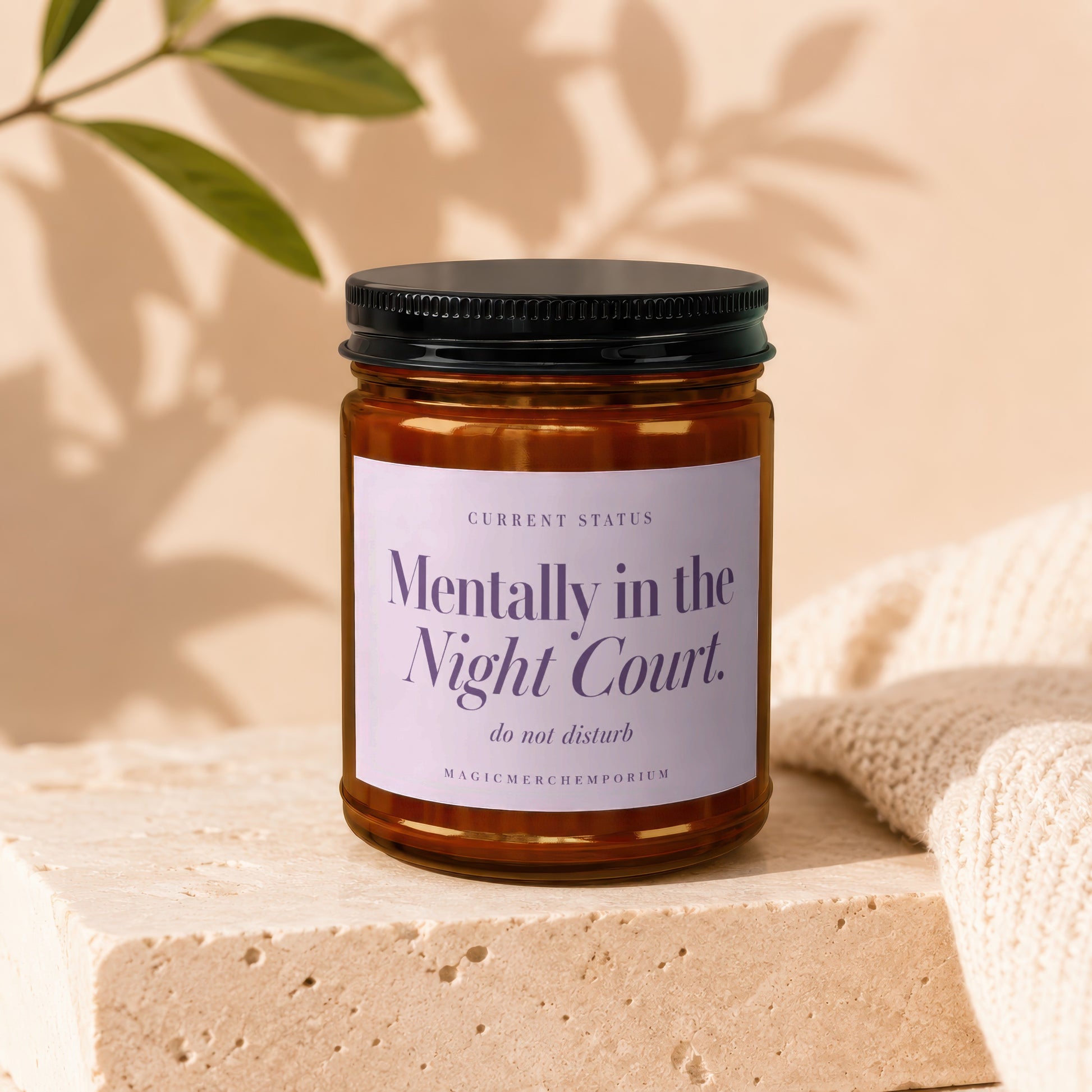 An amber glass candle jar with a black lid and a label reading Mentally in the Night Court sits on a stone block.