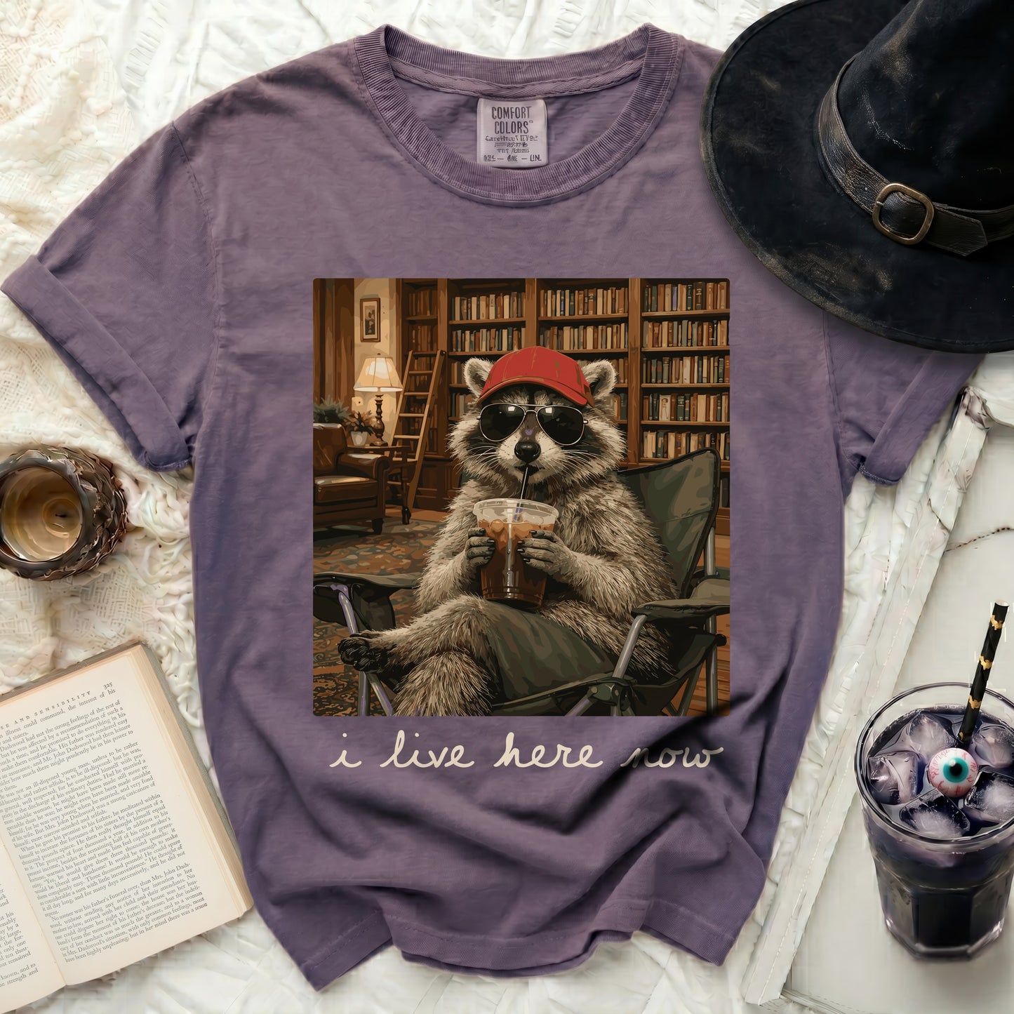 Purple t-shirt with a graphic of a raccoon in sunglasses and a hat, drinking coffee, with text "i live here now".
