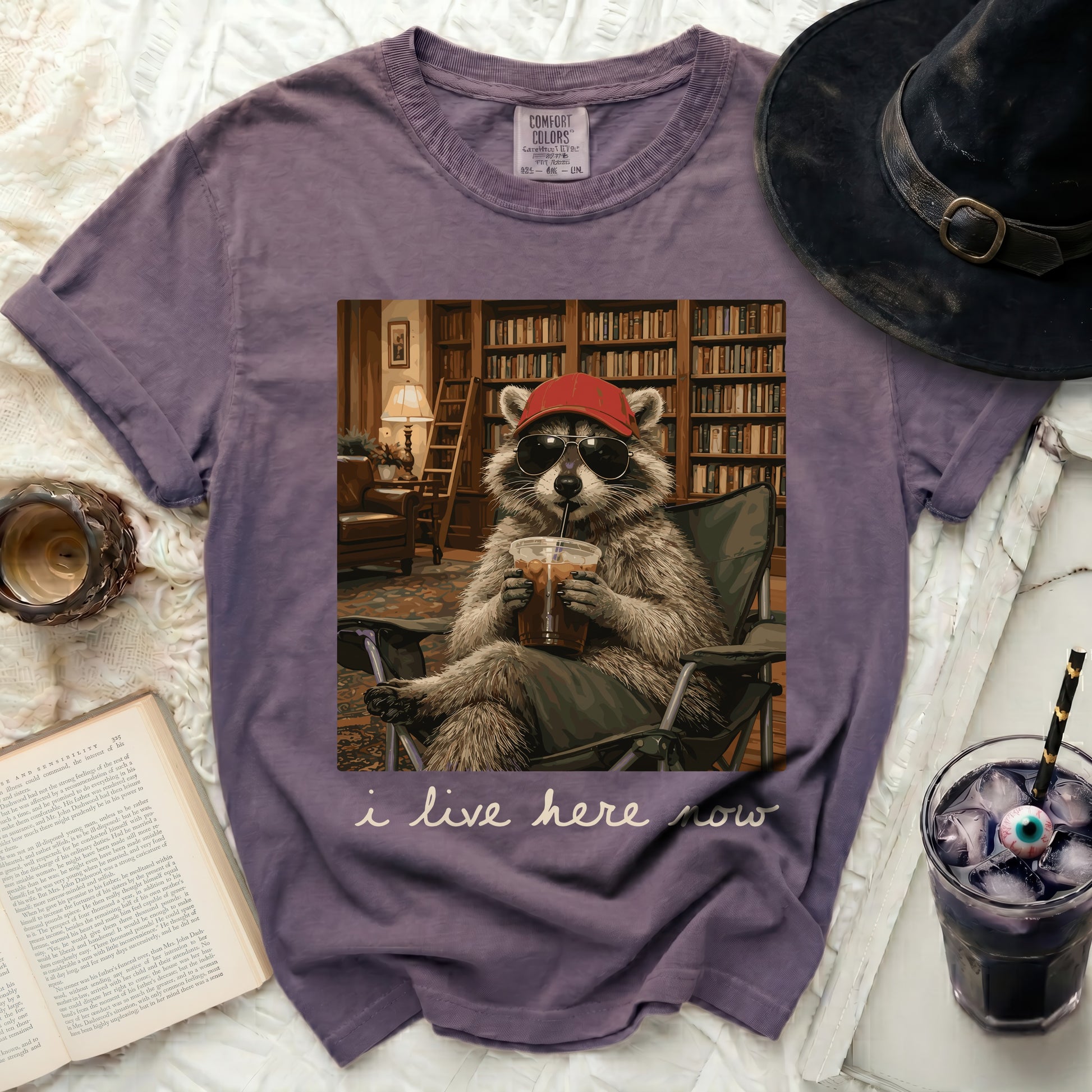 Purple t-shirt with a graphic of a raccoon in sunglasses and a hat, drinking coffee, with text "i live here now".