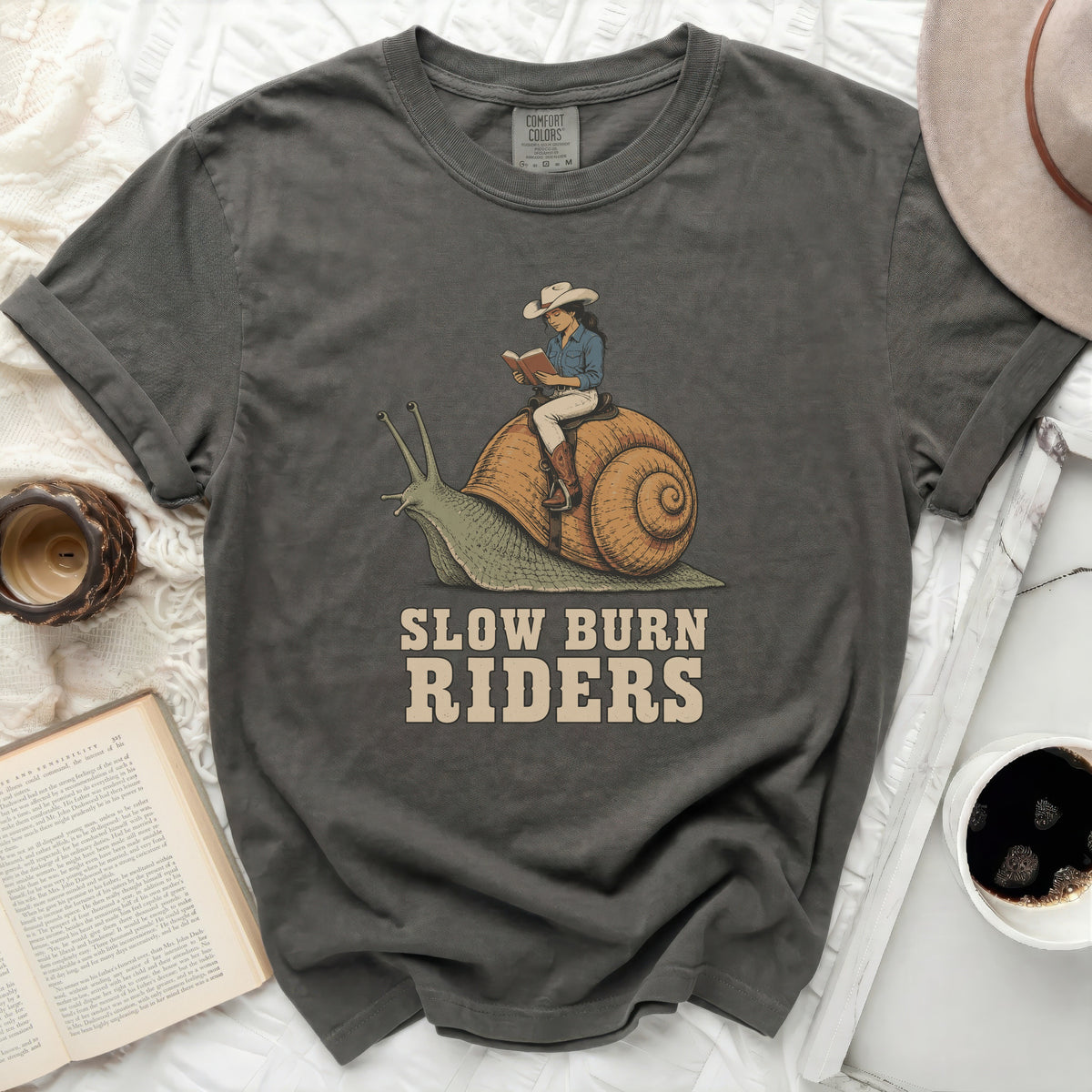 T-shirt graphic with cowgirl reading on a snail and text "SLOW BURN RIDERS".