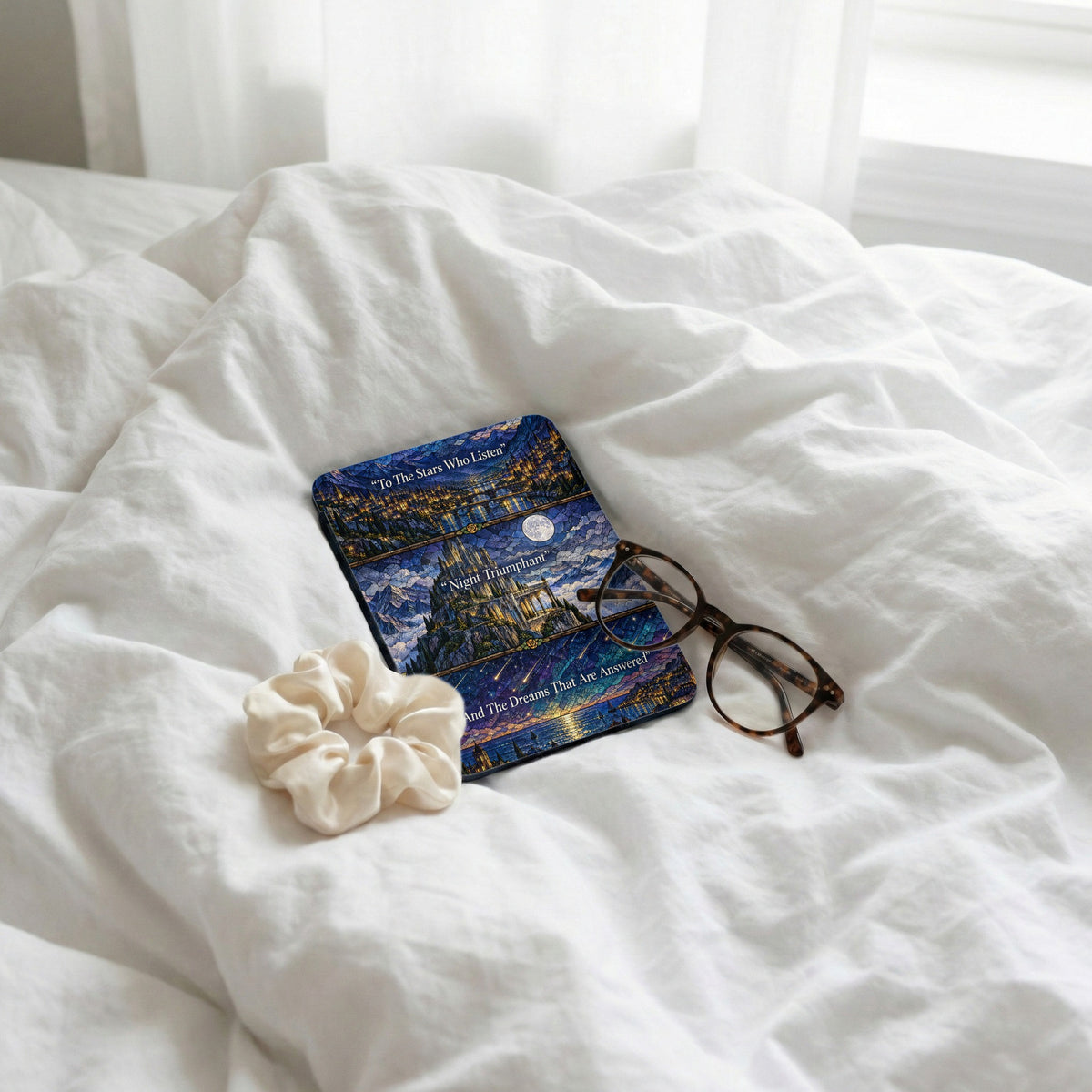 A Kindle case with a starry night castle design lies on a white bed with glasses and a scrunchie.