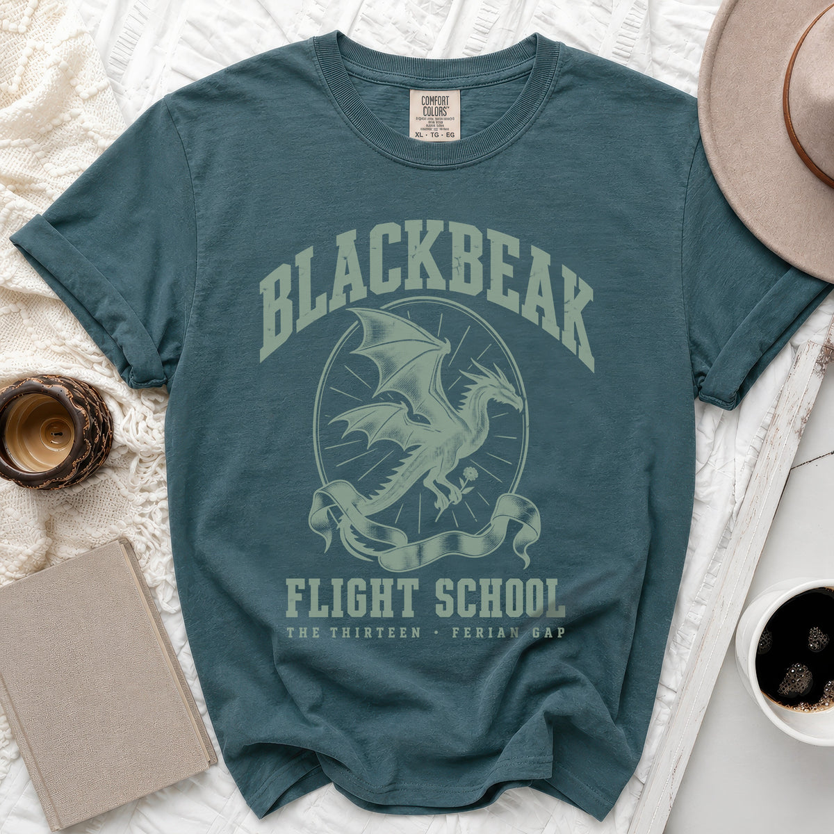 A blue-green t-shirt features a vintage dragon graphic, the text "BLACKBEAK FLIGHT SCHOOL," and surrounding props.