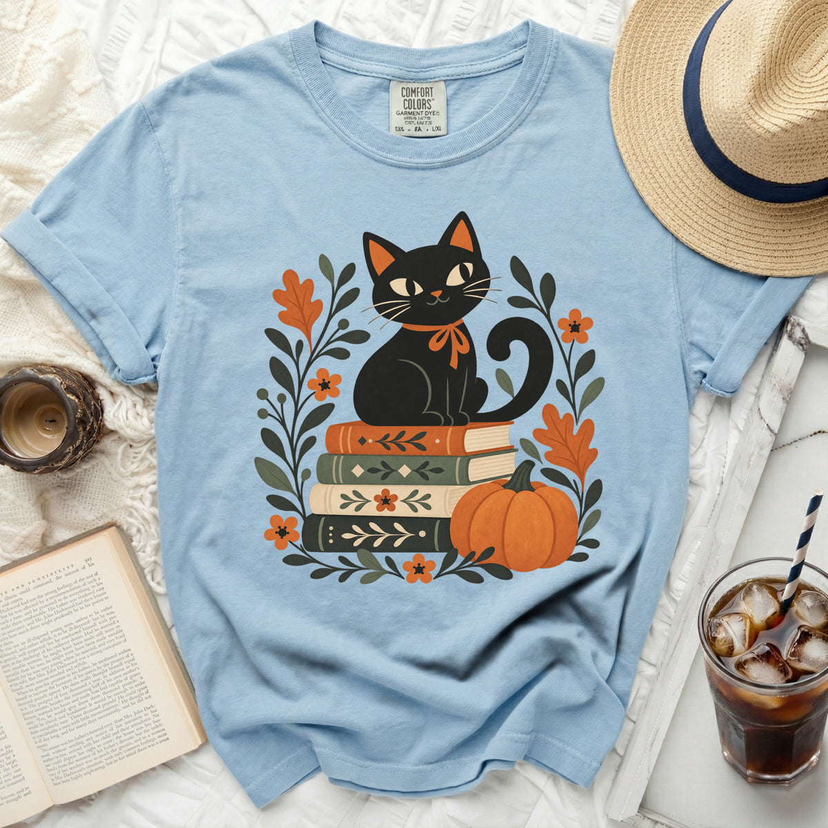 Light blue t-shirt with a black cat sitting on books surrounded by fall leaves.