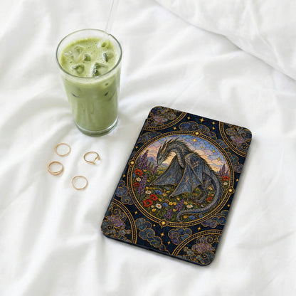 An e-reader with an ornate dragon case rests on white bedding beside an iced matcha latte and gold rings.