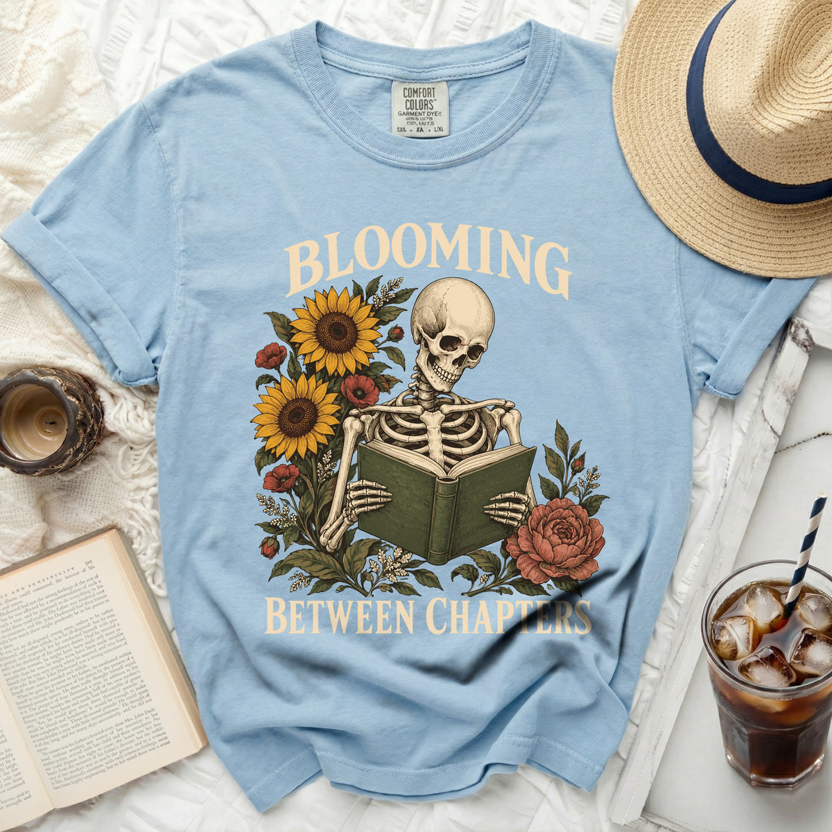 Light blue t-shirt with skeleton reading a book, sunflowers, and roses, text "BLOOMING BETWEEN CHAPTERS".