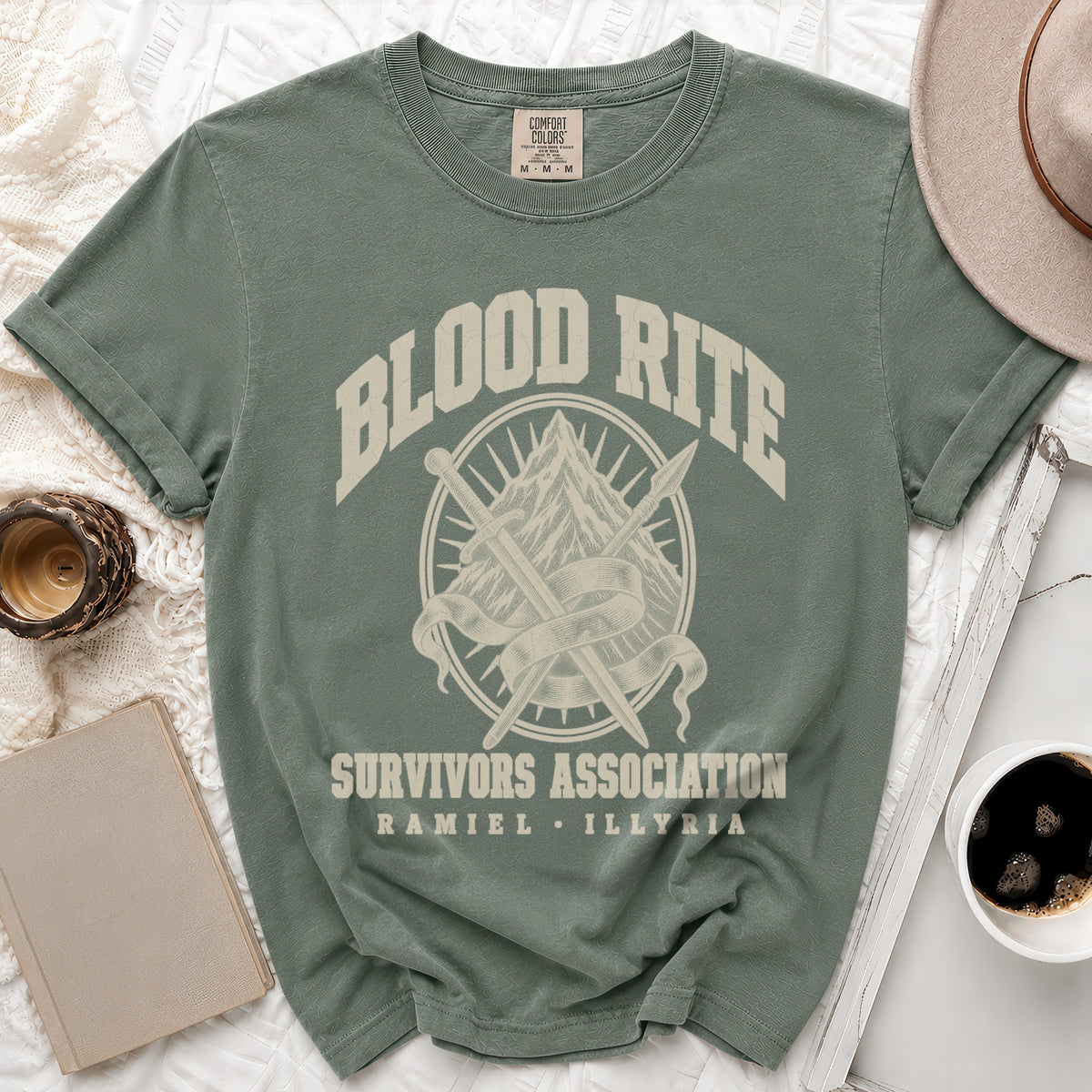 A muted green short-sleeve t-shirt with a graphic of crossed swords, a mountain, and text reading "Blood Rite Survivors Association Ramiel Illyria".