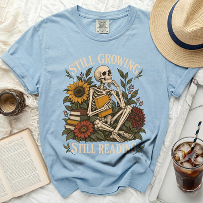 Light blue t-shirt with skeleton, sunflowers, flowers, books, and text: "Still Growing Still Reading".