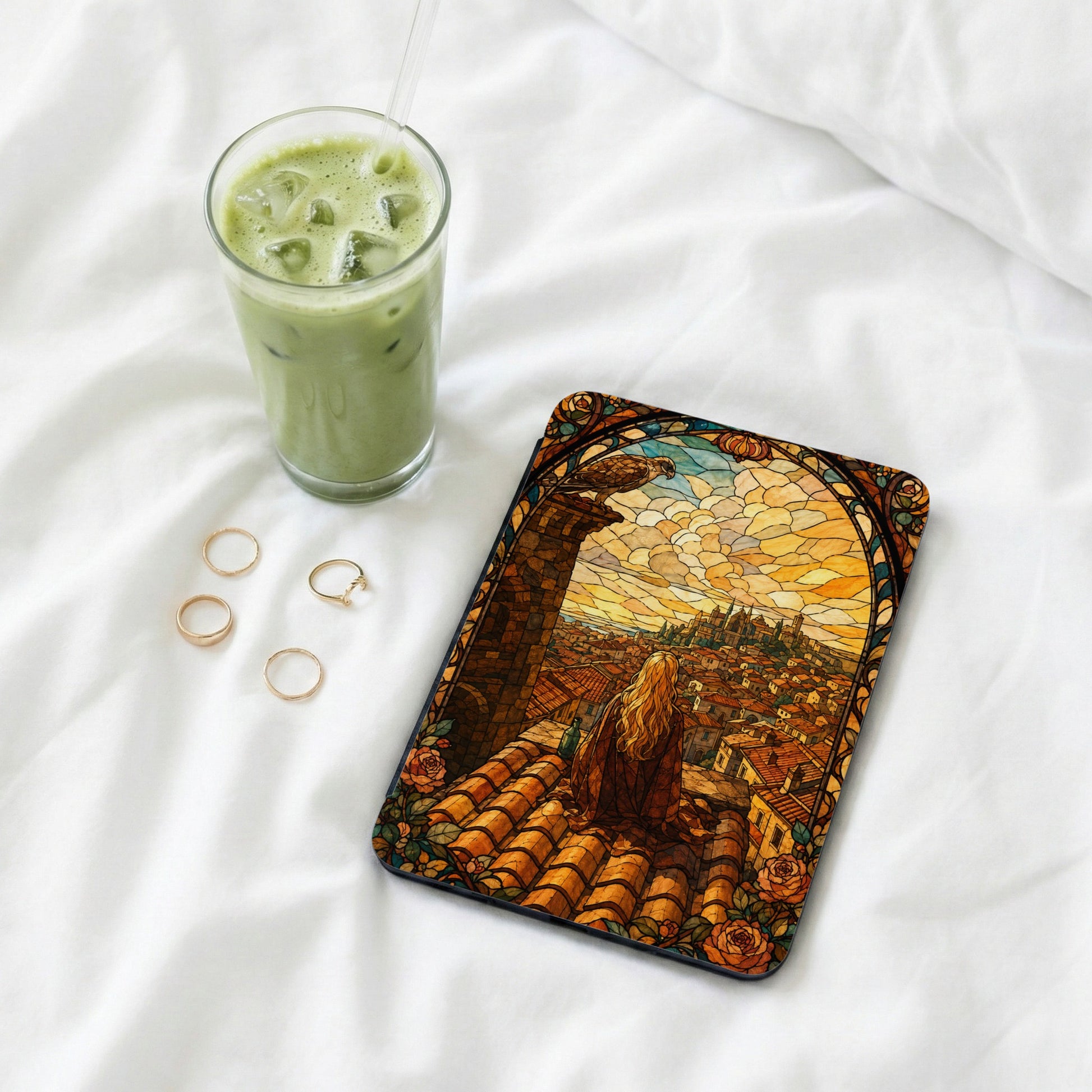 Tablet case with stained glass art of woman and city, next to drink and rings.