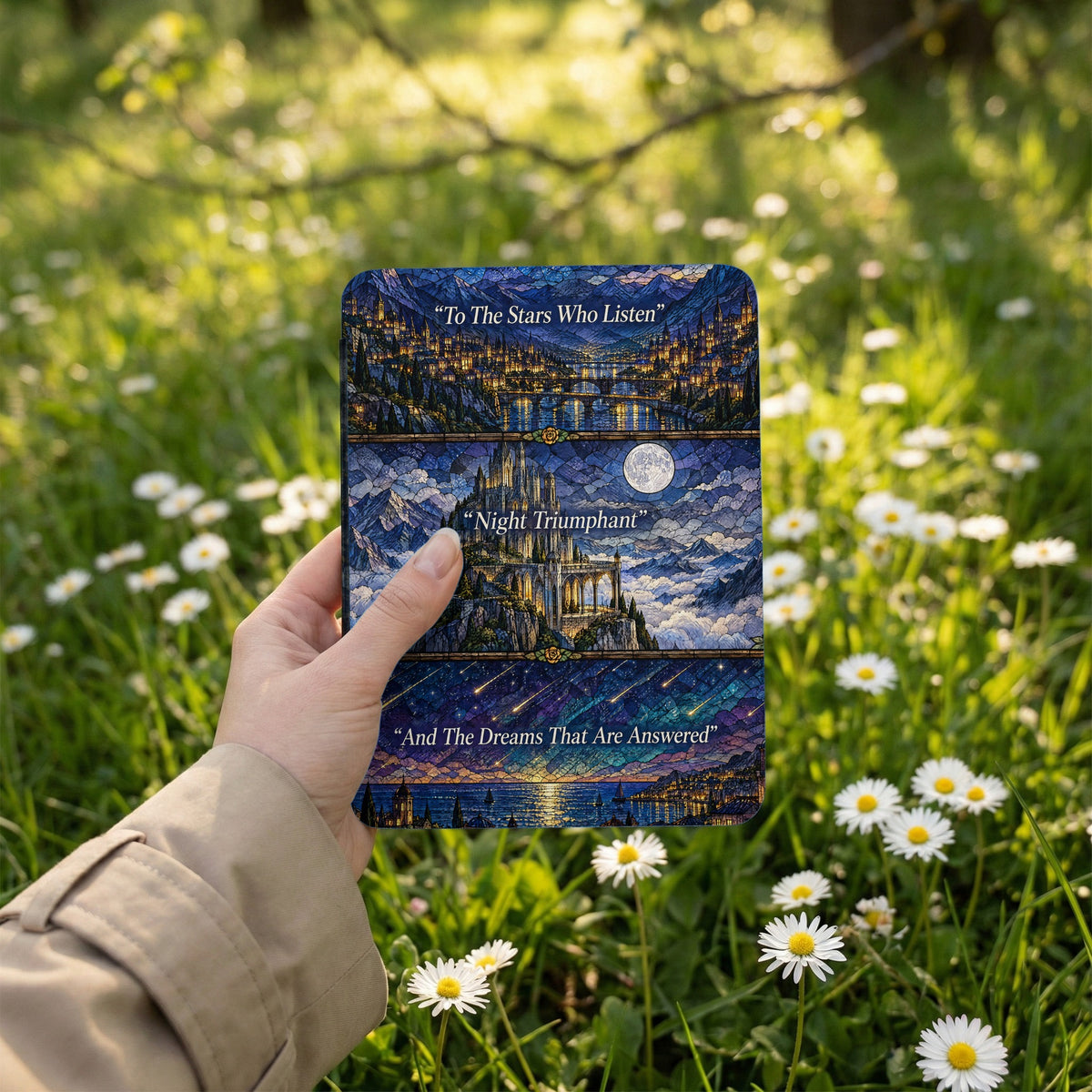 A hand holds an e-reader decorated with a fantasy stained-glass design against a grassy, daisy-filled meadow.
