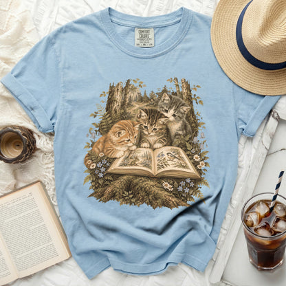 Light blue t-shirt with three kittens reading a book in a forest scene.