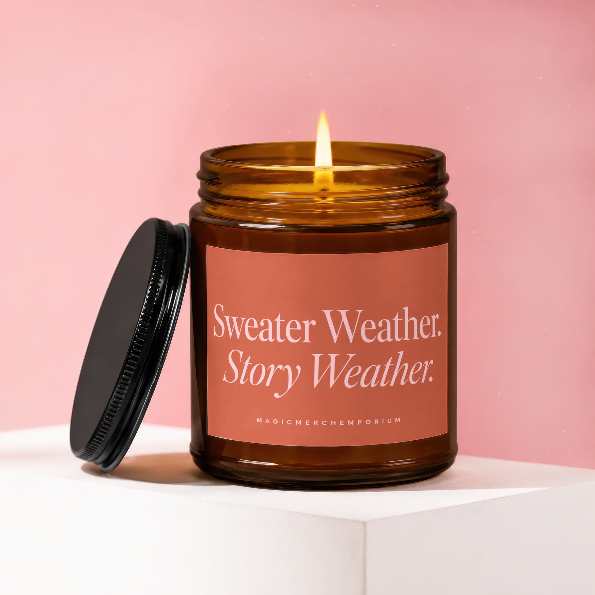 Amber jar candle with lit flame and coral label reading "Sweater Weather. Story Weather."