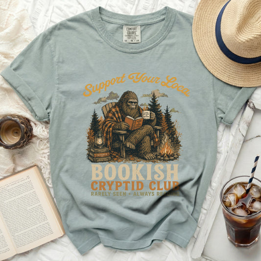 Bigfoot reads a book and drinks coffee in a forest with "Bookish Cryptid Club" text.