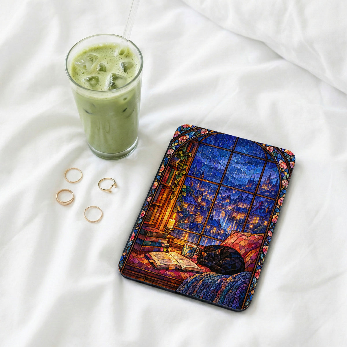 An e-reader with a stained-glass style case lies on white sheets beside four rings and an iced matcha latte.