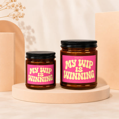 Two amber glass candles with pink labels reading "My WIP Is Winning" on display.