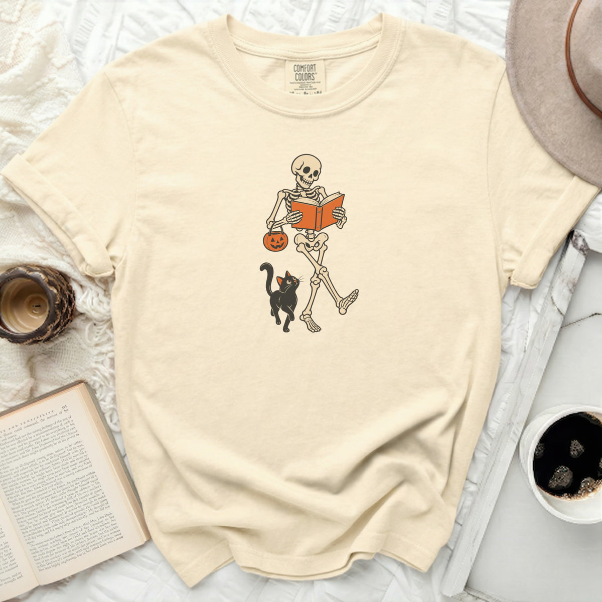 Skeleton reading book with pumpkin bucket and cat on cream t-shirt.
