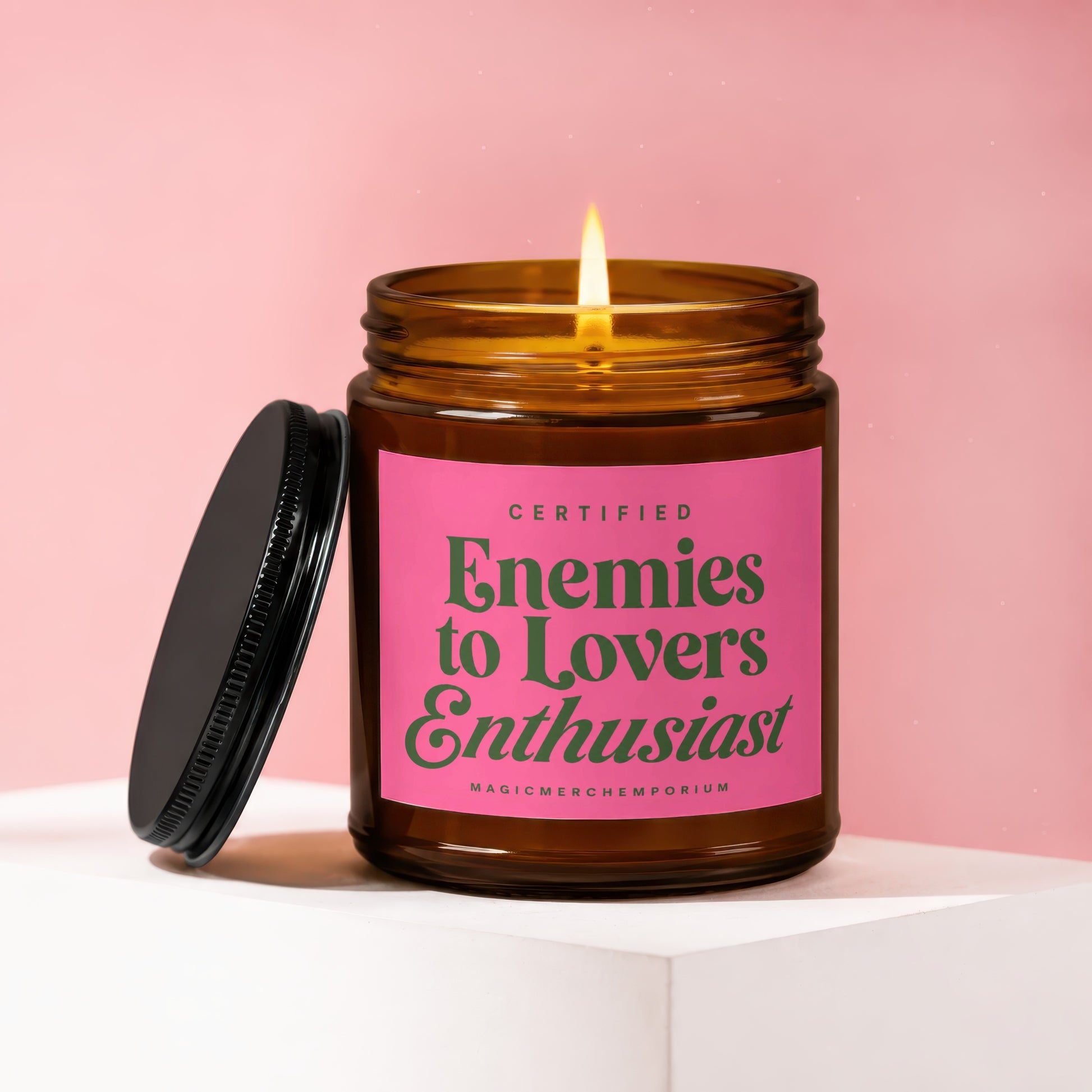 Amber jar candle with lit flame, pink label reading "Certified Enemies to Lovers Enthusiast".