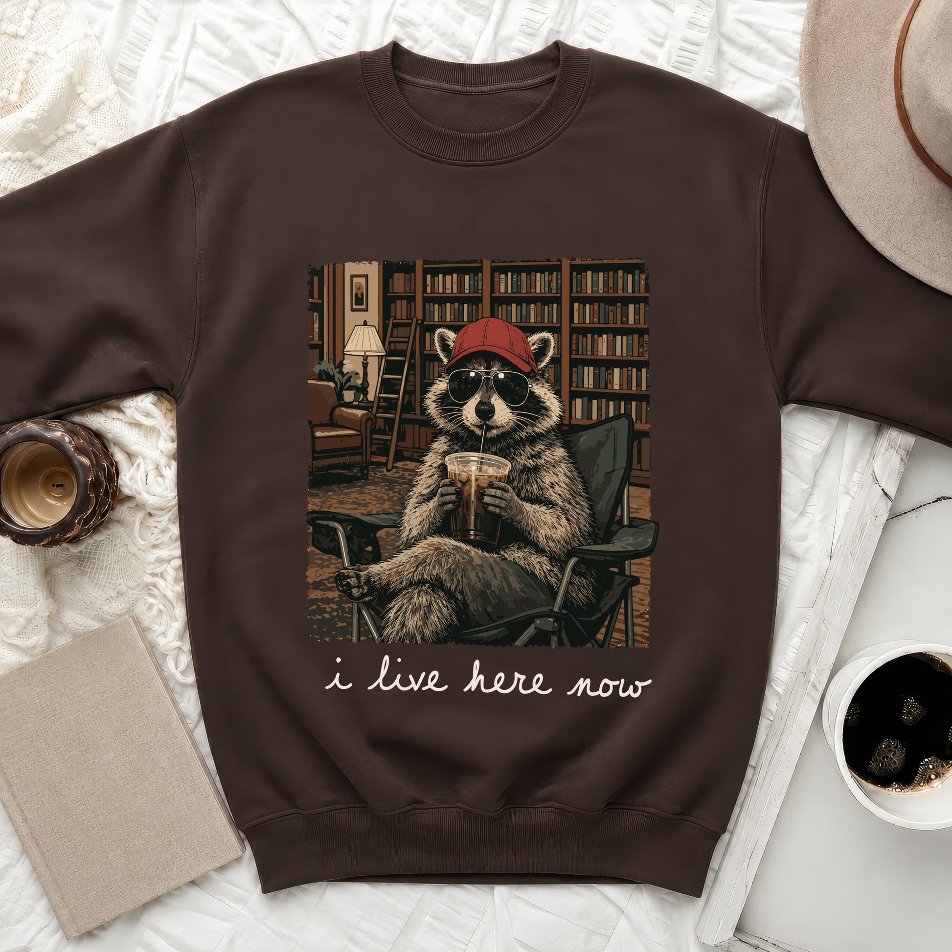 Brown sweatshirt with graphic of raccoon reading in library, drinking coffee, and text "i live here now".
