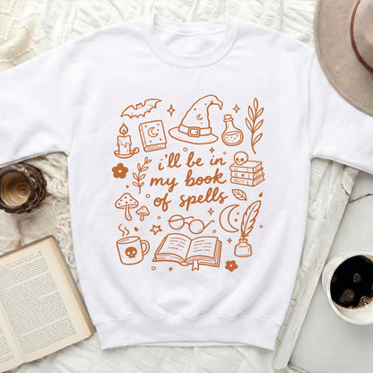 White sweatshirt with "I'll be in my book of spells" and whimsical illustrations.