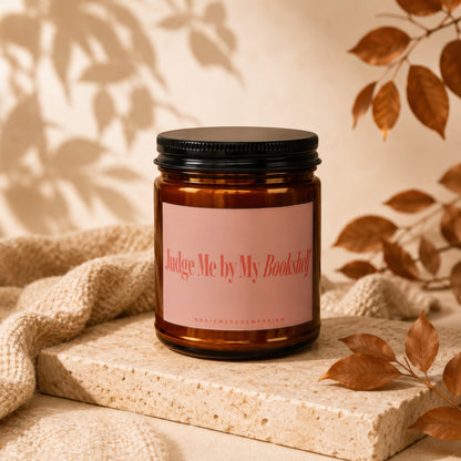 Amber jar candle with pink label reading "Judge Me by My Bookshelf."