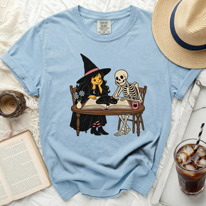 Light blue t-shirt with a cartoon of a witch and skeleton reading.