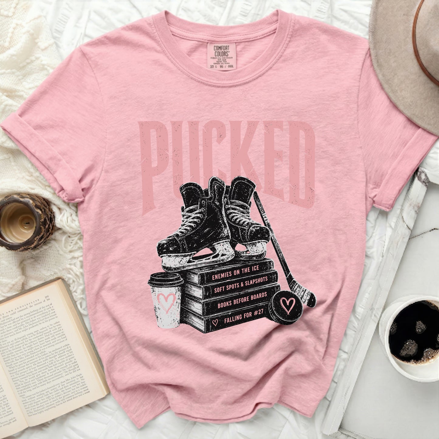 Pink t-shirt with "Pucked" graphic over hockey skates, books, and coffee cup.