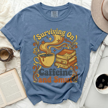Blue t-shirt with graphic of coffee cup, books, and text "Surviving On Caffeine and Smut".